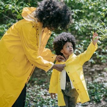 A mother helps her child button a yellow raincoat while exploring a lush green forest.
